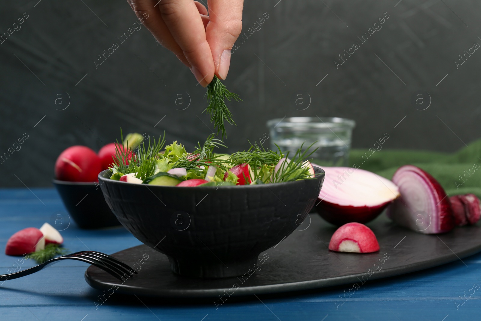 Photo of Woman adding dill into bowl of salad with radish at blue wooden table, closeup