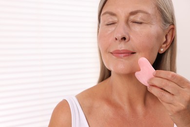 Woman massaging her face with rose quartz gua sha tool in bathroom, closeup. Space for text