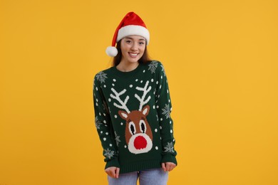 Photo of Happy young woman in Santa hat showing Christmas sweater on orange background