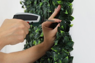 Photo of Man with screwdriver installing green artificial plant panel on white wall, closeup