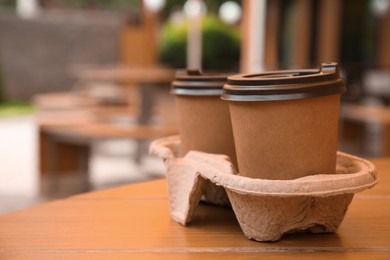 Takeaway paper coffee cups with plastic lids in cardboard holder on wooden table outdoors, space for text