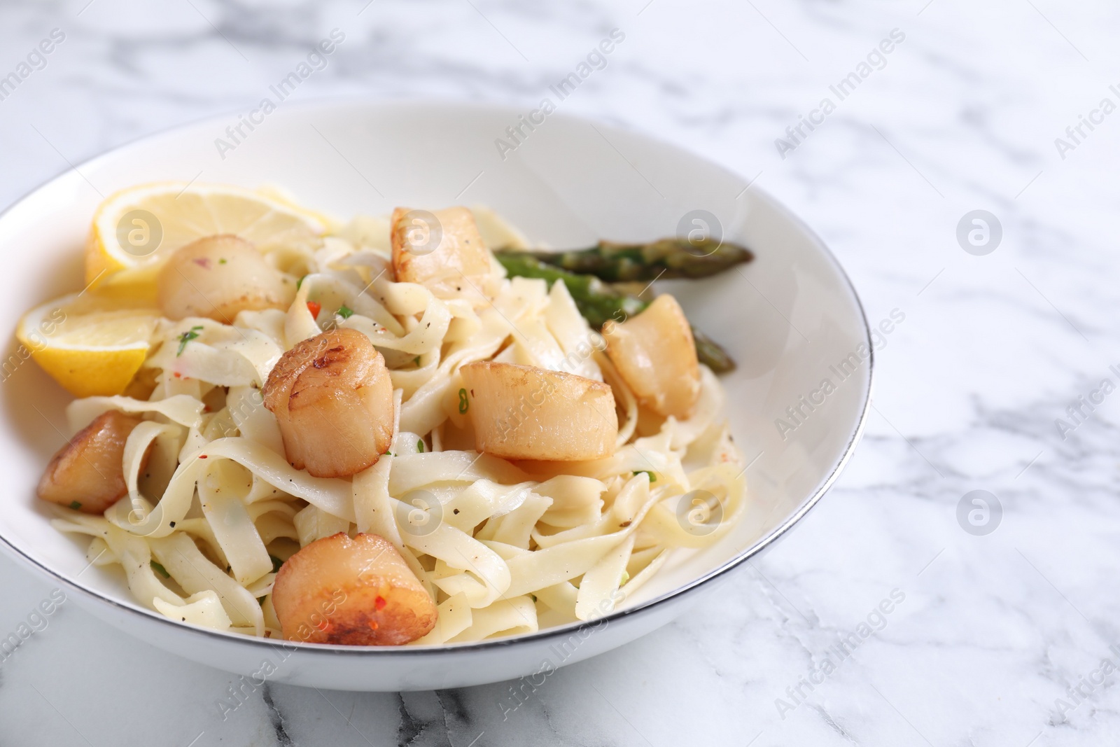Photo of Delicious scallop pasta with asparagus and lemon on white marble table, closeup