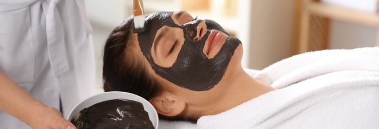 Cosmetologist applying black mask onto woman's face in spa salon. Banner design