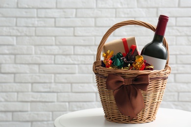 Photo of Wicker gift basket with food and wine on table near white brick wall. Space for text