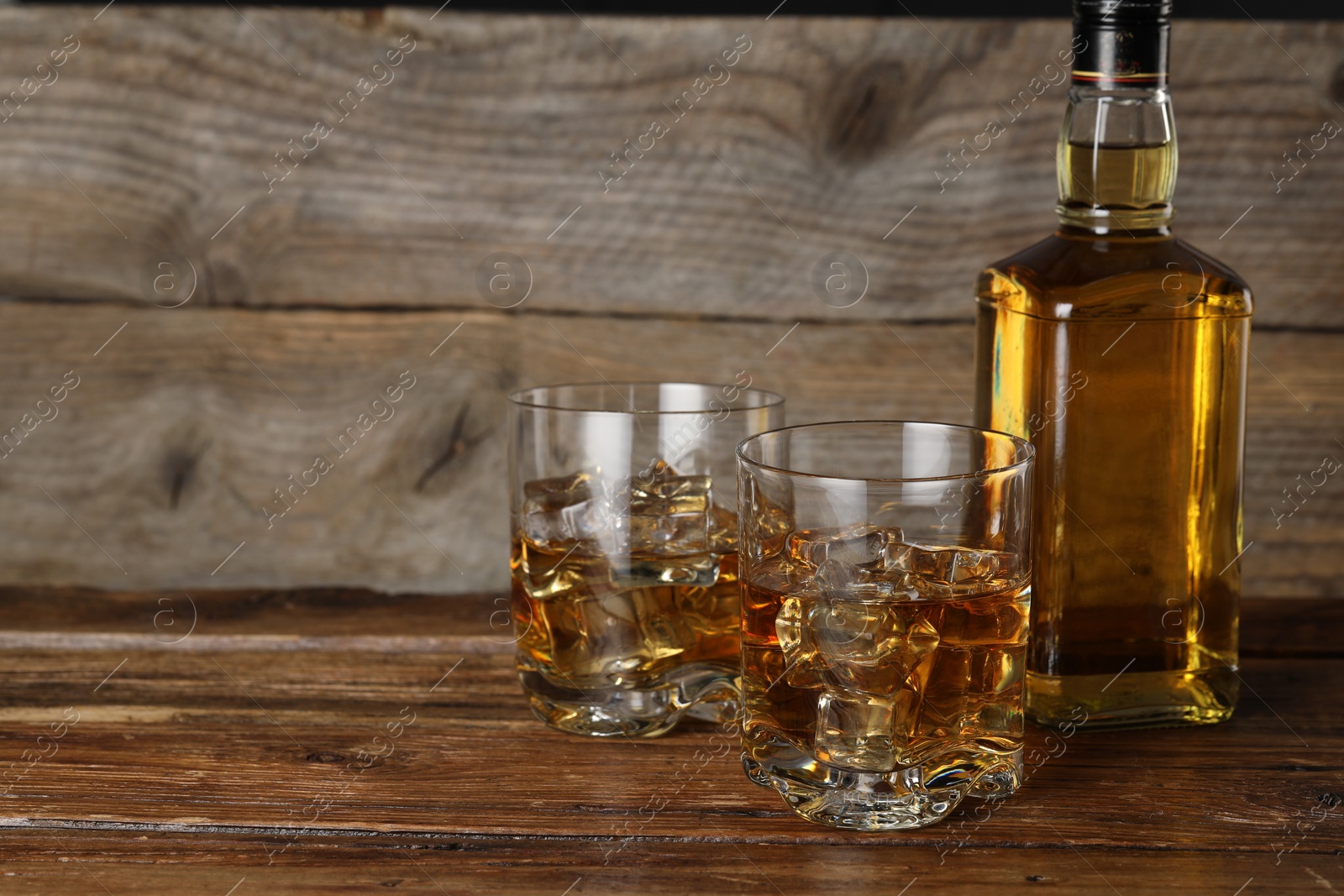 Photo of Whiskey with ice cubes in glasses and bottle on wooden table, closeup. Space for text