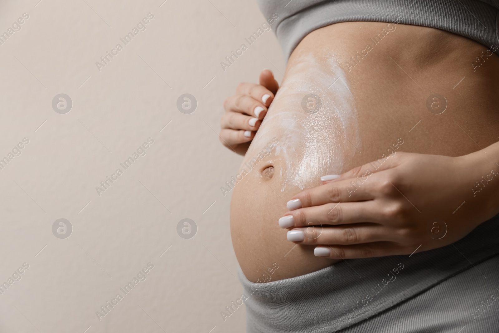 Photo of Pregnant woman applying cosmetic product on belly against beige background, closeup. Space for text