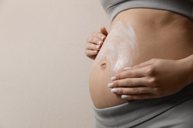 Pregnant woman applying cosmetic product on belly against beige background, closeup. Space for text
