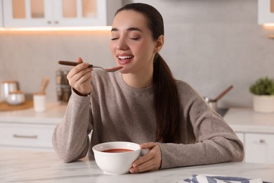 Photo of Smiling woman eating tasty soup at white marble table in kitchen