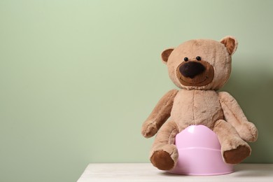 Photo of Teddy bear on pink baby potty on white wooden table against olive background, space for text. Toilet training