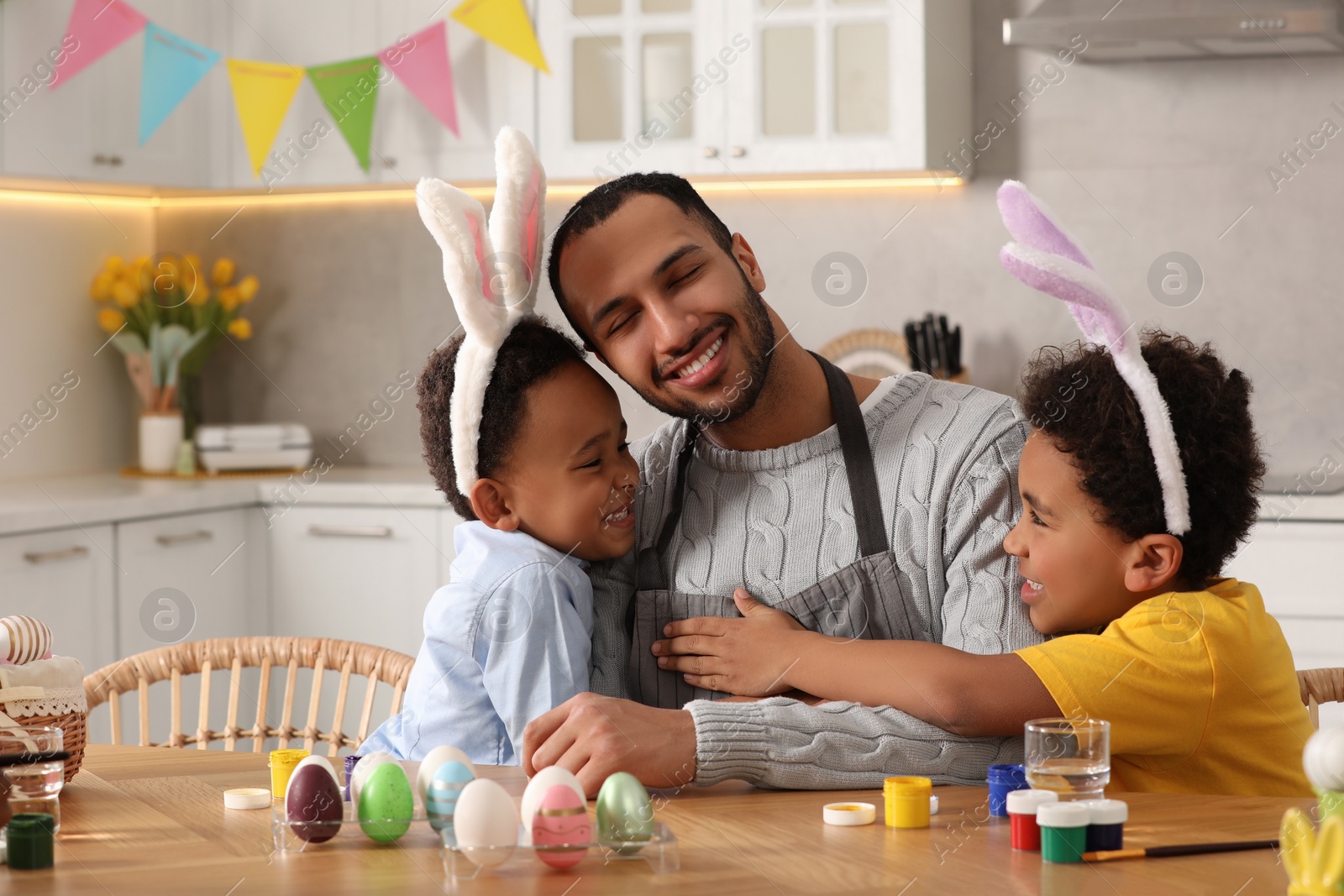 Photo of Happy African American father hugging his cute children and Easter eggs at table in kitchen