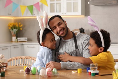 Happy African American father hugging his cute children and Easter eggs at table in kitchen