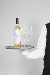 Young waiter holding tray with glasses and bottle of wine on light background