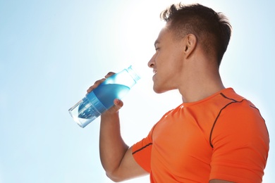 Young sporty man drinking water from bottle against blue sky on sunny day. Space for text
