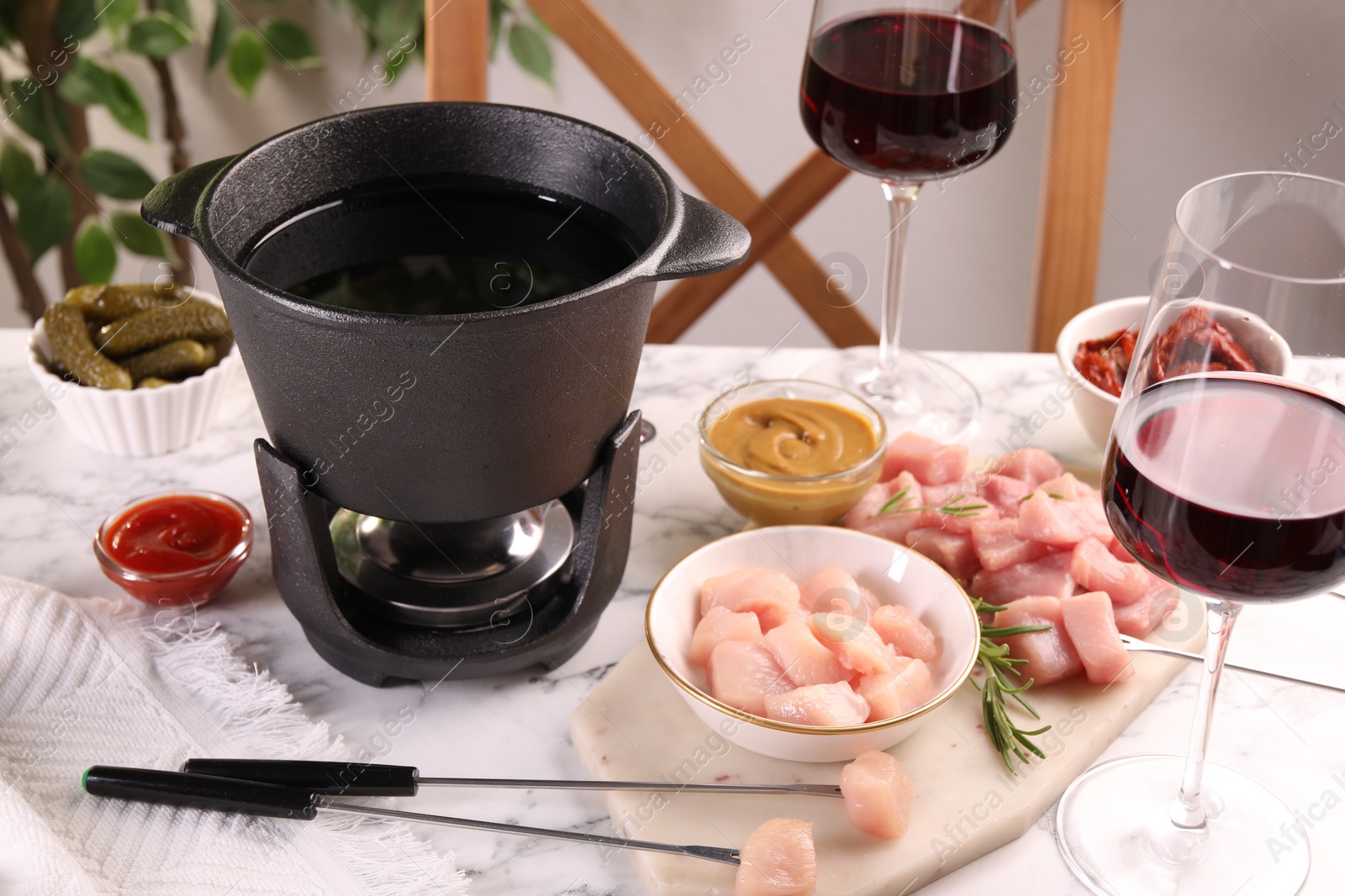 Photo of Fondue pot with oil, forks, raw meat pieces, glasses of red wine and other products on white marble table