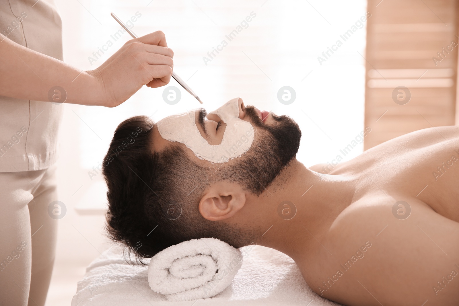 Photo of Cosmetologist applying mask on man's face in spa salon