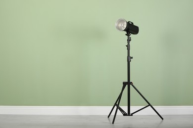 Studio flash light with reflector on tripod near pale green wall in room, space for text. Professional photographer's equipment
