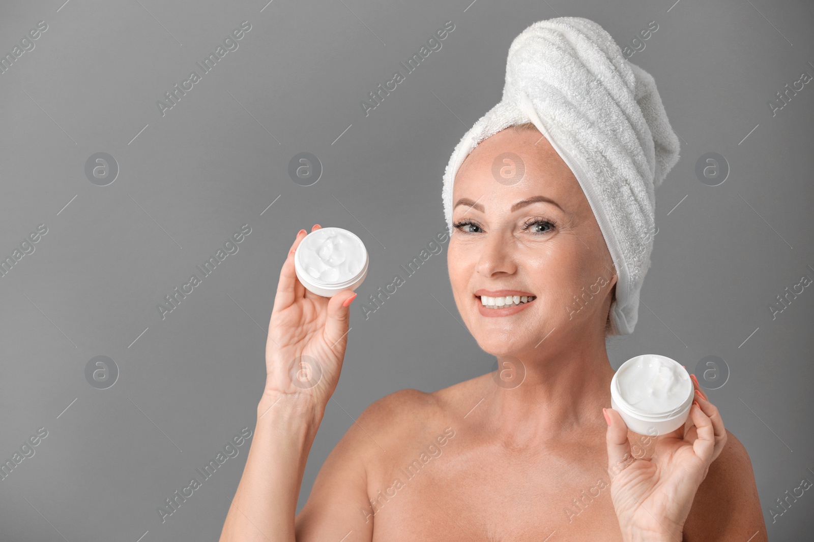 Photo of Portrait of beautiful mature woman with perfect skin holding jars of cream on grey background
