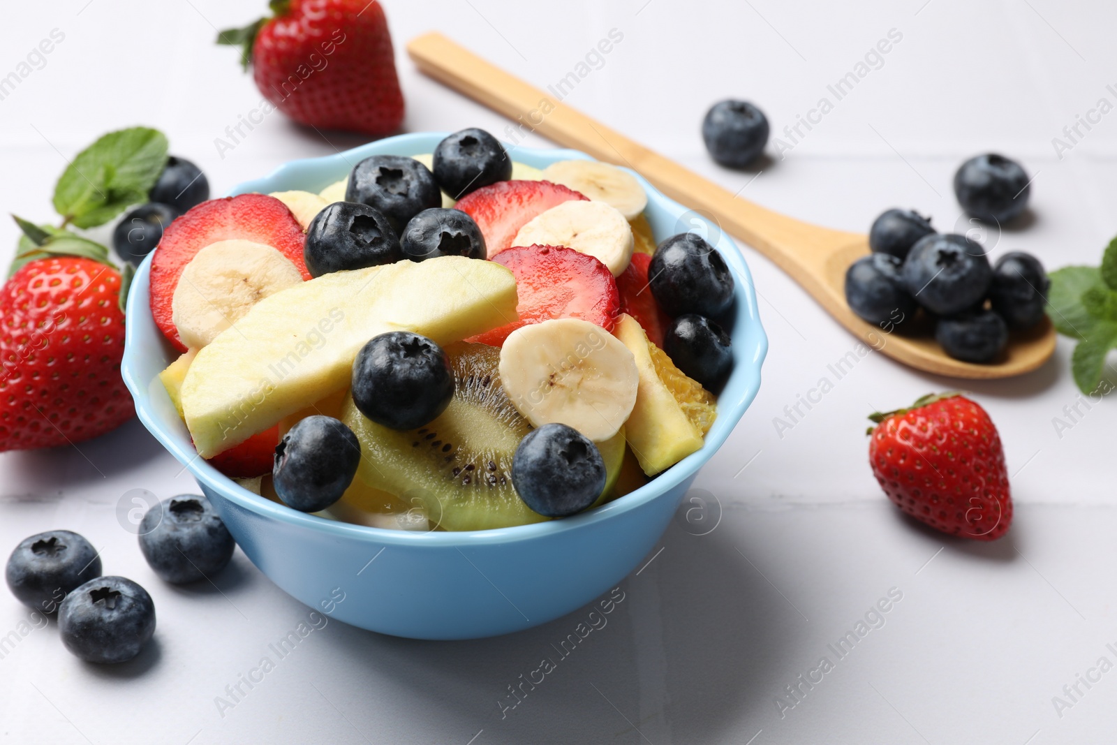 Photo of Tasty fruit salad in bowl and ingredients on white tiled table, closeup
