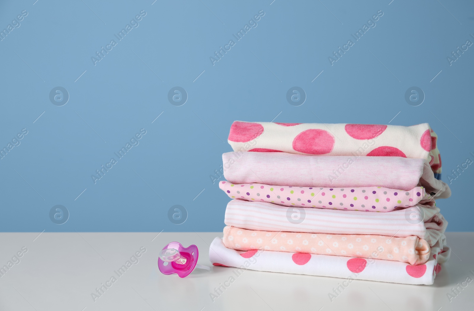 Photo of Stack of stylish child clothes and pacifier on table against color background