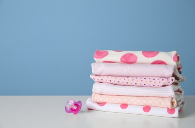 Stack of stylish child clothes and pacifier on table against color background