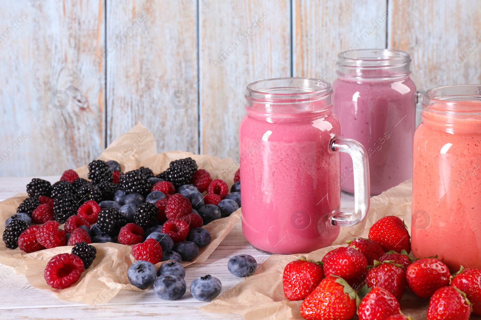 Photo of Mason jars of different berry smoothies and fresh ingredients on white wooden table