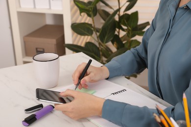 Photo of Woman taking notes while using smartphone at white marble table indoors, closeup