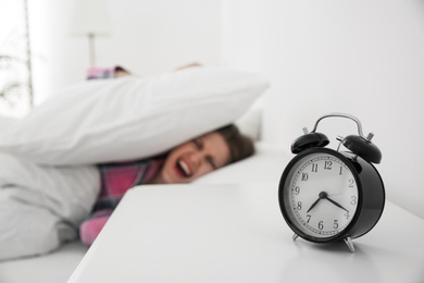 Young woman covering ears with pillows at home in morning, focus on alarm clock