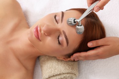 Photo of Young woman receiving facial massage with metal roller in beauty salon, above view