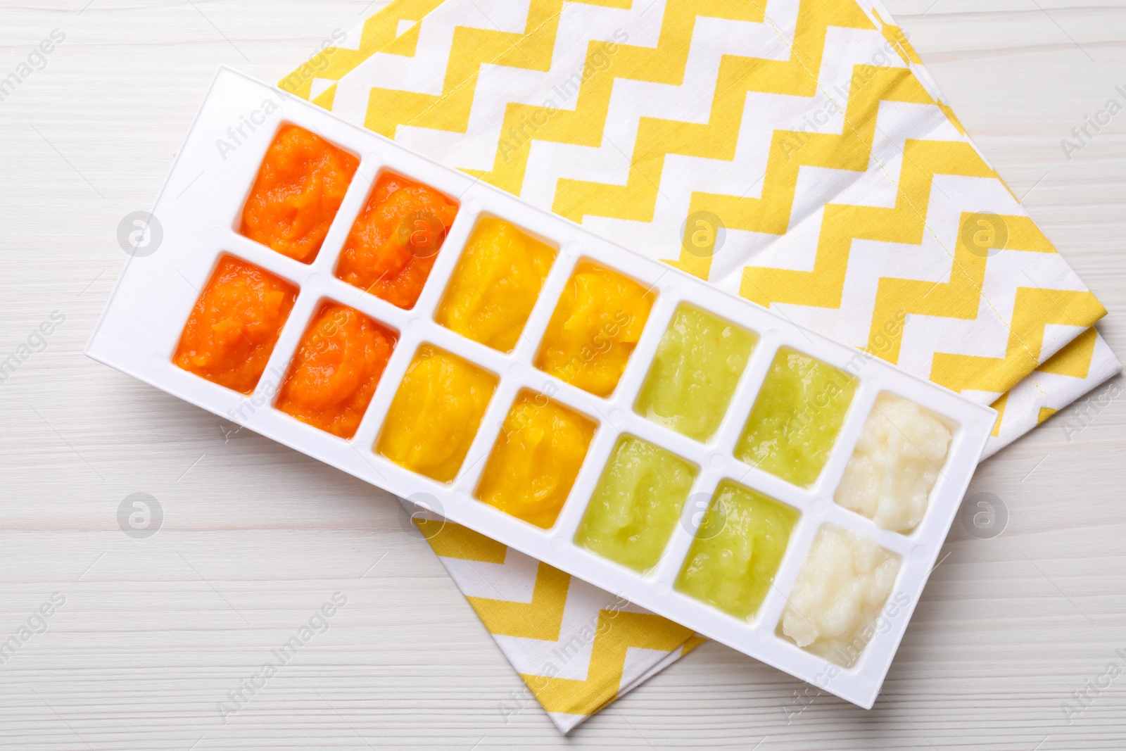 Photo of Different purees in ice cube tray on white wooden table, top view. Ready for freezing