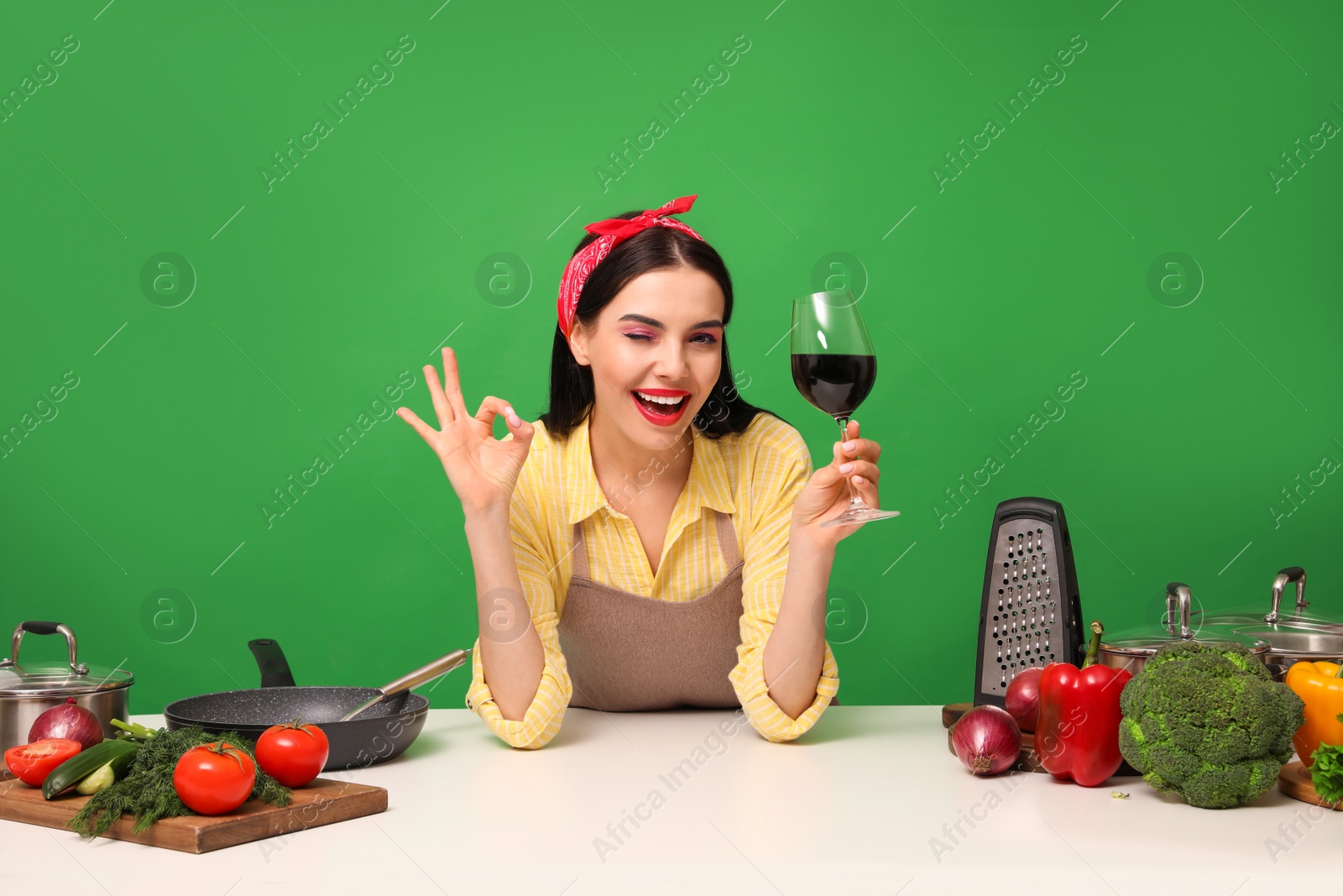 Photo of Young housewife with glass of wine, vegetables and different utensils on green background