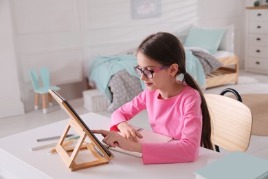 Photo of Little girl doing homework with tablet at table in bedroom