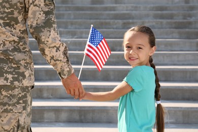 Photo of Soldier and his little daughter with American flag holding hands outdoors. Veterans Day in USA