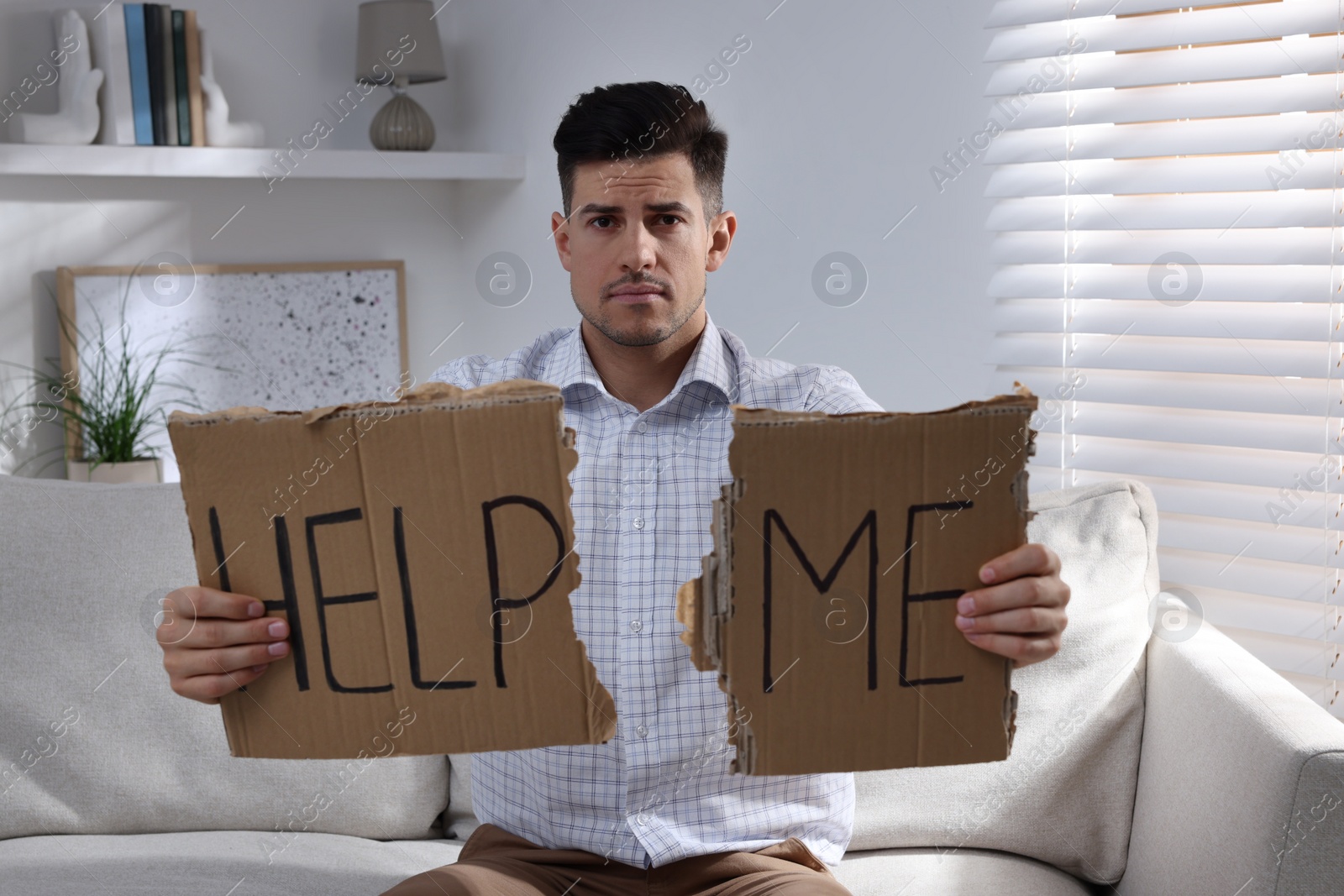 Photo of Unhappy man with HELP ME sign on sofa indoors