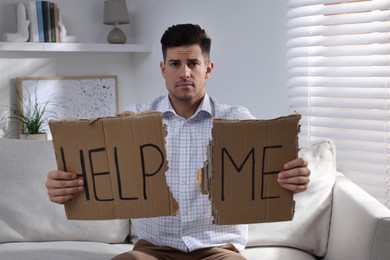 Unhappy man with HELP ME sign on sofa indoors