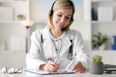 Photo of Smiling doctor in headphones having online consultation and filling documents at table indoors