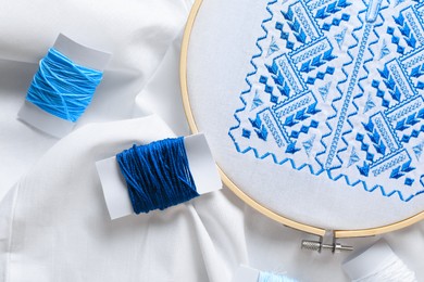 Photo of Threads and blue Ukrainian national embroidery on white fabric, top view