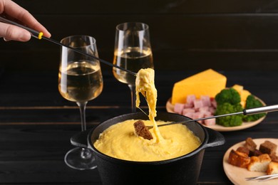 Woman dipping pieces of ham and bread into fondue pot with melted cheese at table, closeup