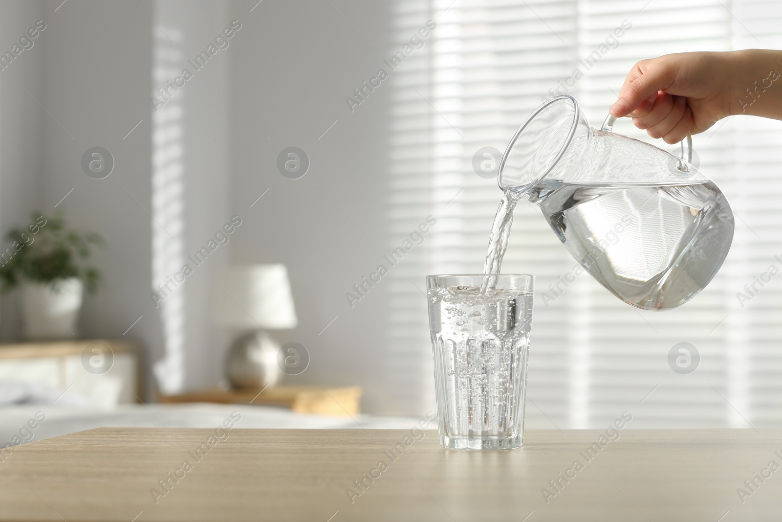 Photo of Woman pouring water from jug into glass at wooden table indoors, closeup. Space for text