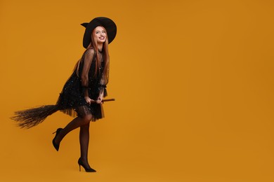 Photo of Happy young woman in scary witch costume with broom posing on orange background, space for text. Halloween celebration