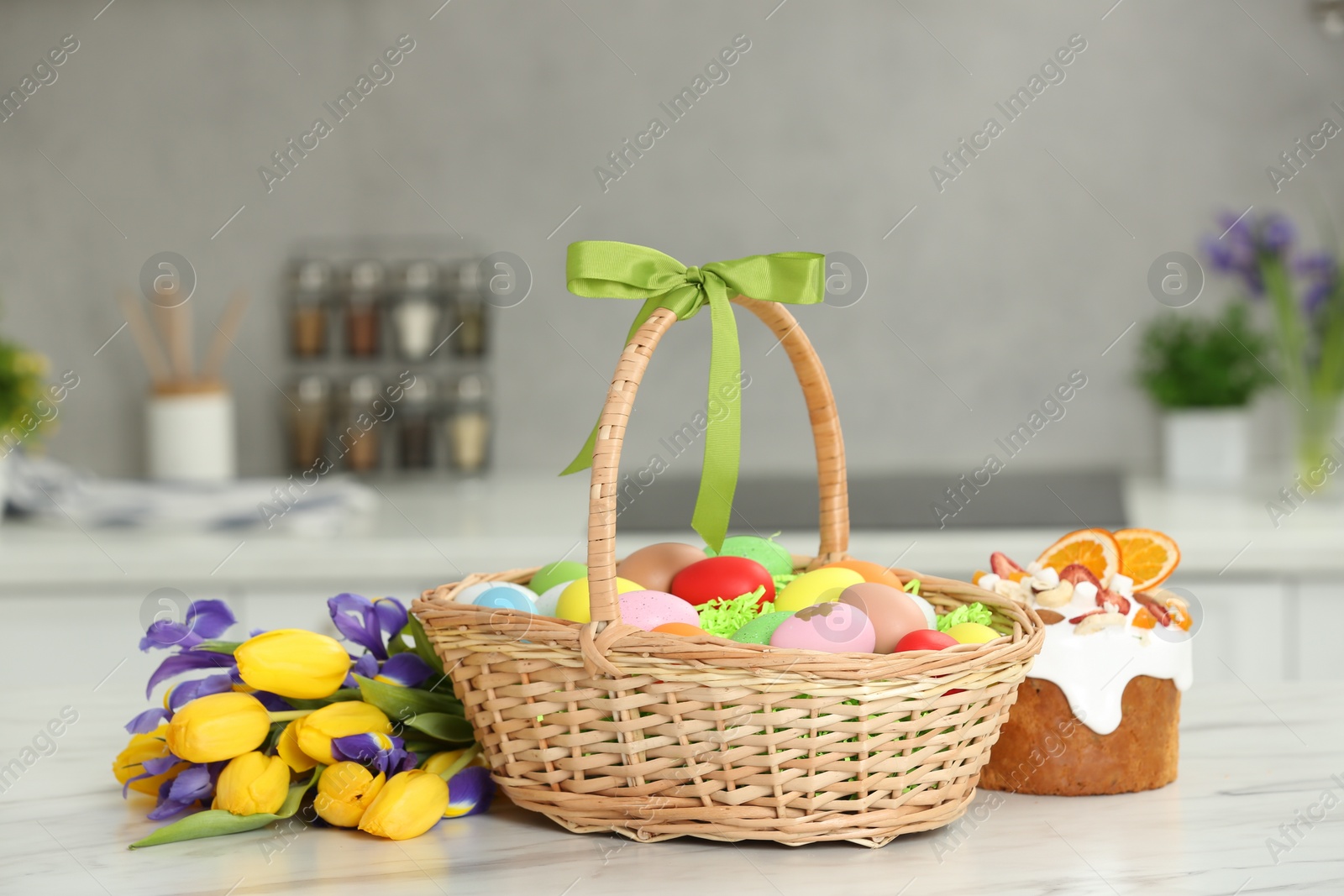 Photo of Easter basket with painted eggs between tulips and cake on white marble table indoors