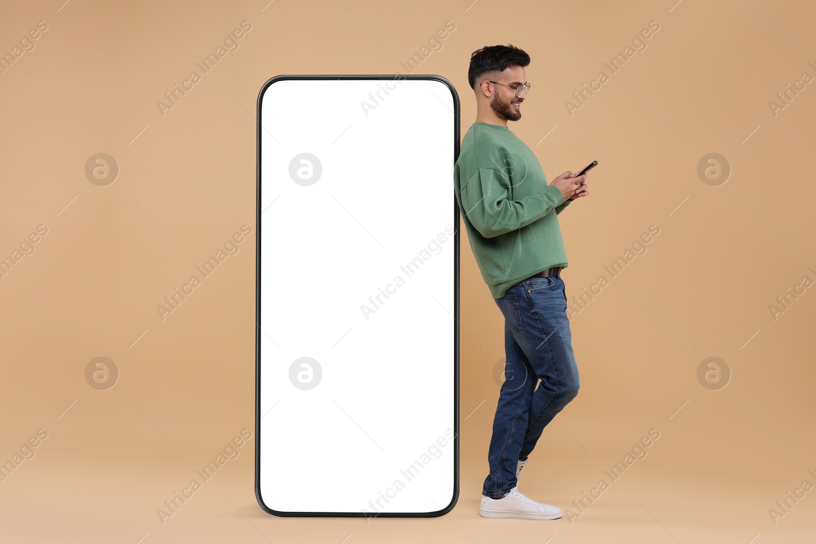 Image of Man with mobile phone standing near huge device with empty screen on dark beige background. Mockup for design