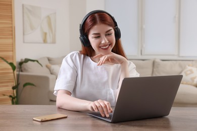 Happy woman with headphones using laptop at wooden table in room