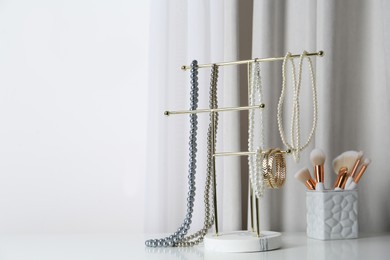 Photo of Interior element. Holder with set of luxurious jewelry and makeup brushes on white dressing table. space for text