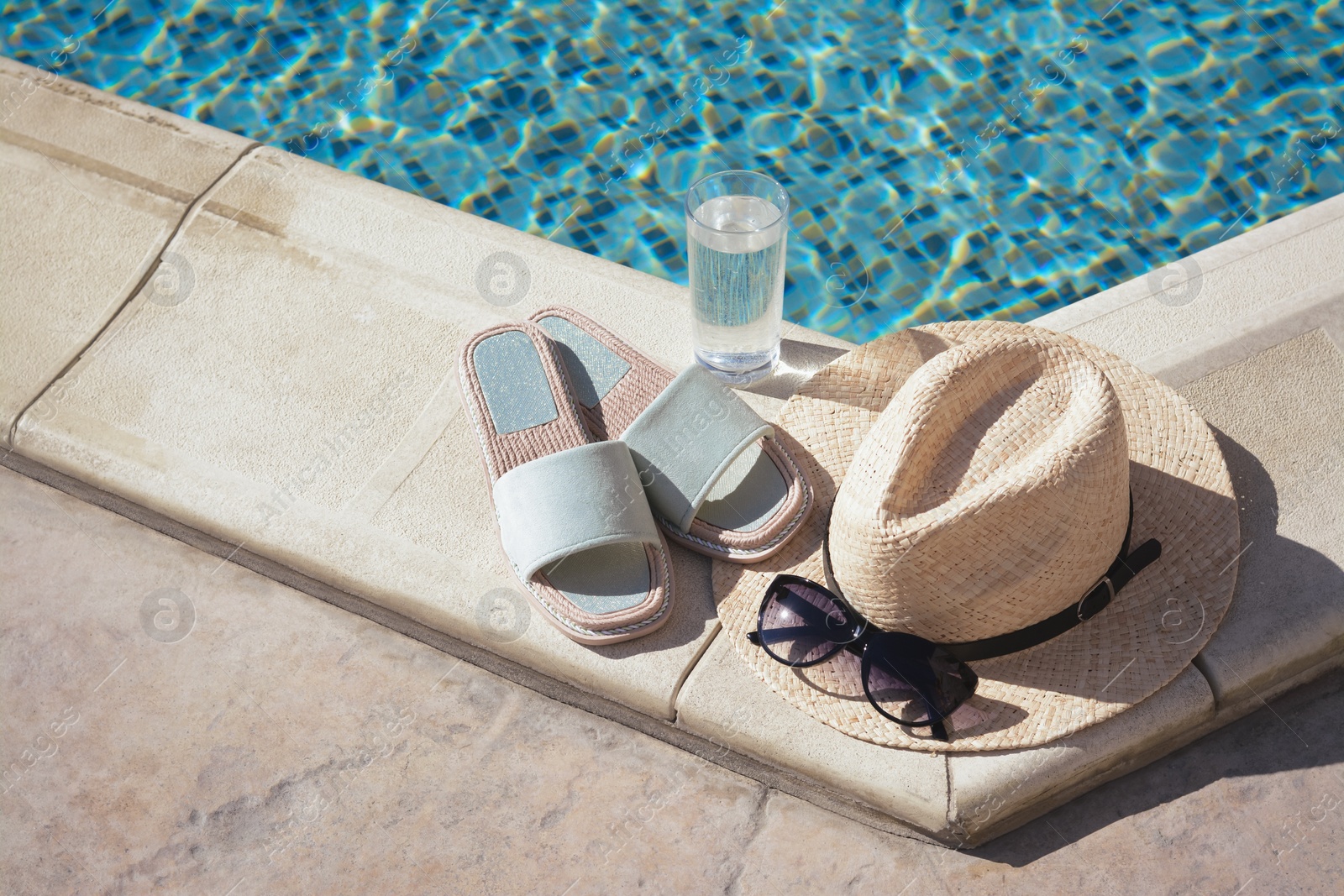 Photo of Stylish sunglasses, slippers, straw hat and glass of water at poolside on sunny day. Beach accessories