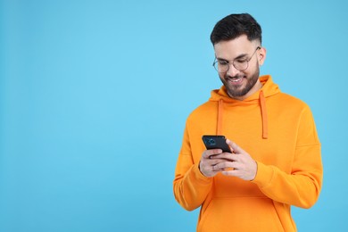 Happy young man using smartphone on light blue background, space for text