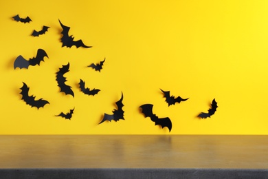 Photo of Empty grey table near yellow wall decorated with paper bats, space for text. Halloween celebration