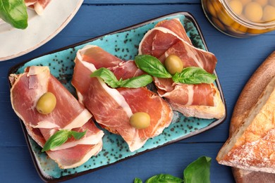 Photo of Tasty sandwiches with cured ham and basil leaves on blue wooden table, flat lay