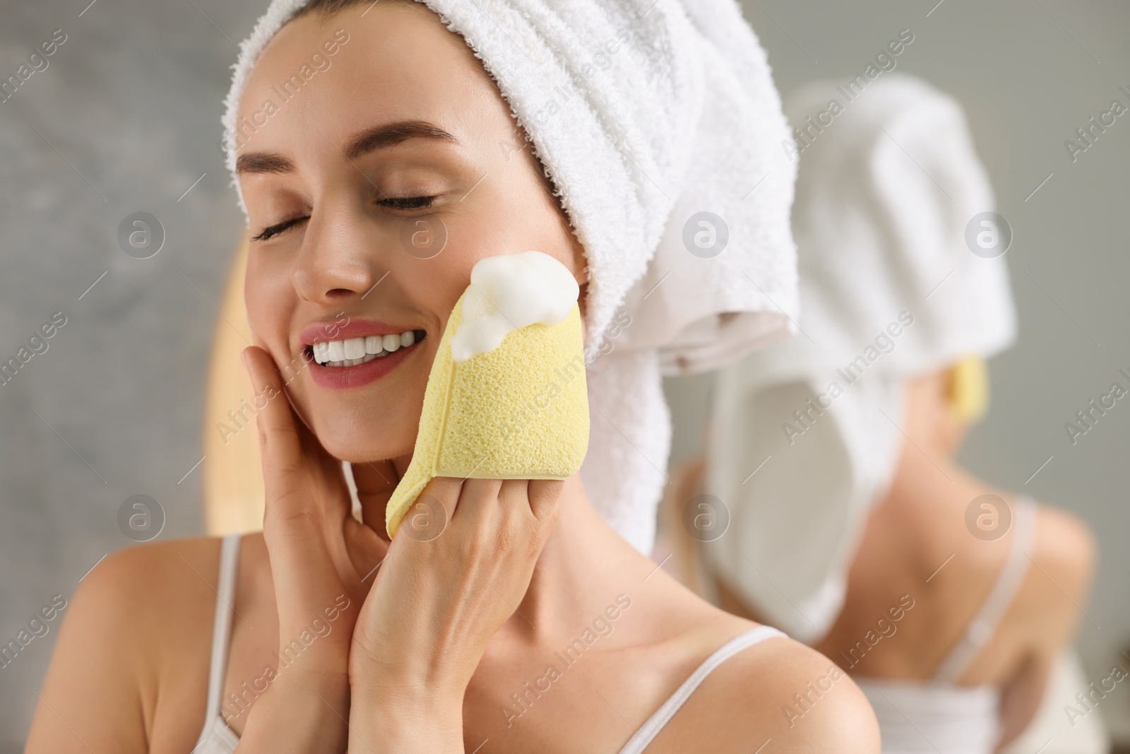 Photo of Happy young woman washing her face with sponge in bathroom