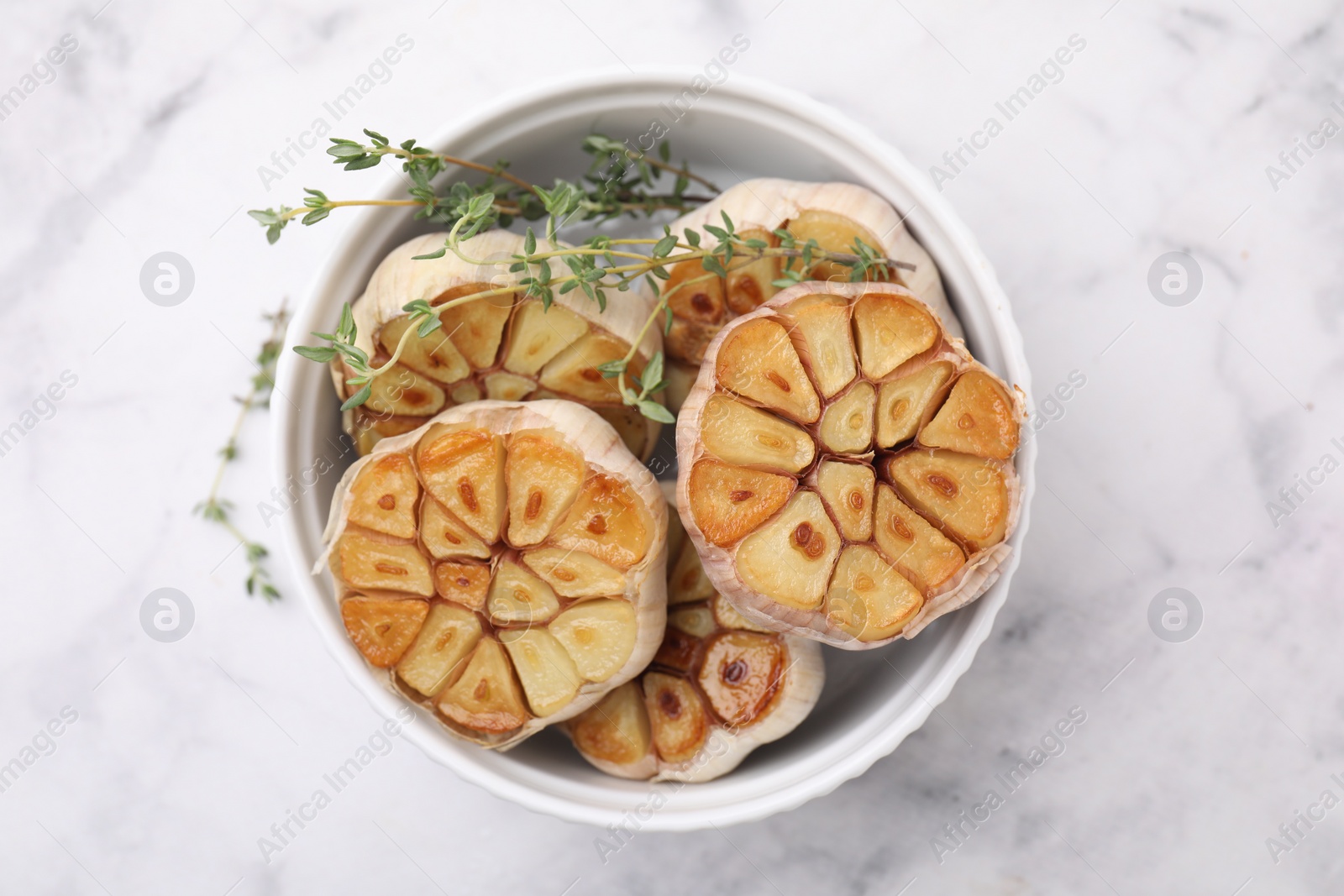 Photo of Heads of fried garlic and thyme in bowl on white table, top view