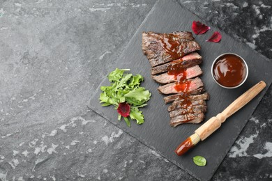 Photo of Pieces of delicious roasted beef meat with sauce and greens on black table, top view. Space for text